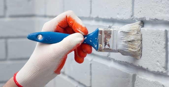 Man in white gloves painting brick wall with white paint brush.